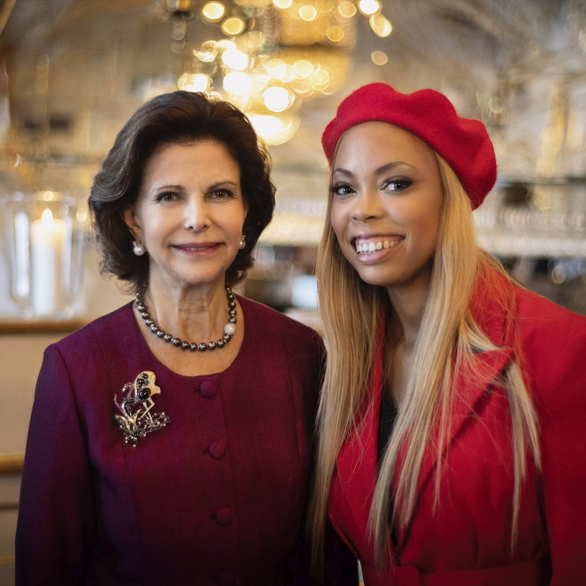 Kiralina standing besides Queen Silvia of Sweden in a red hat and blazer.
