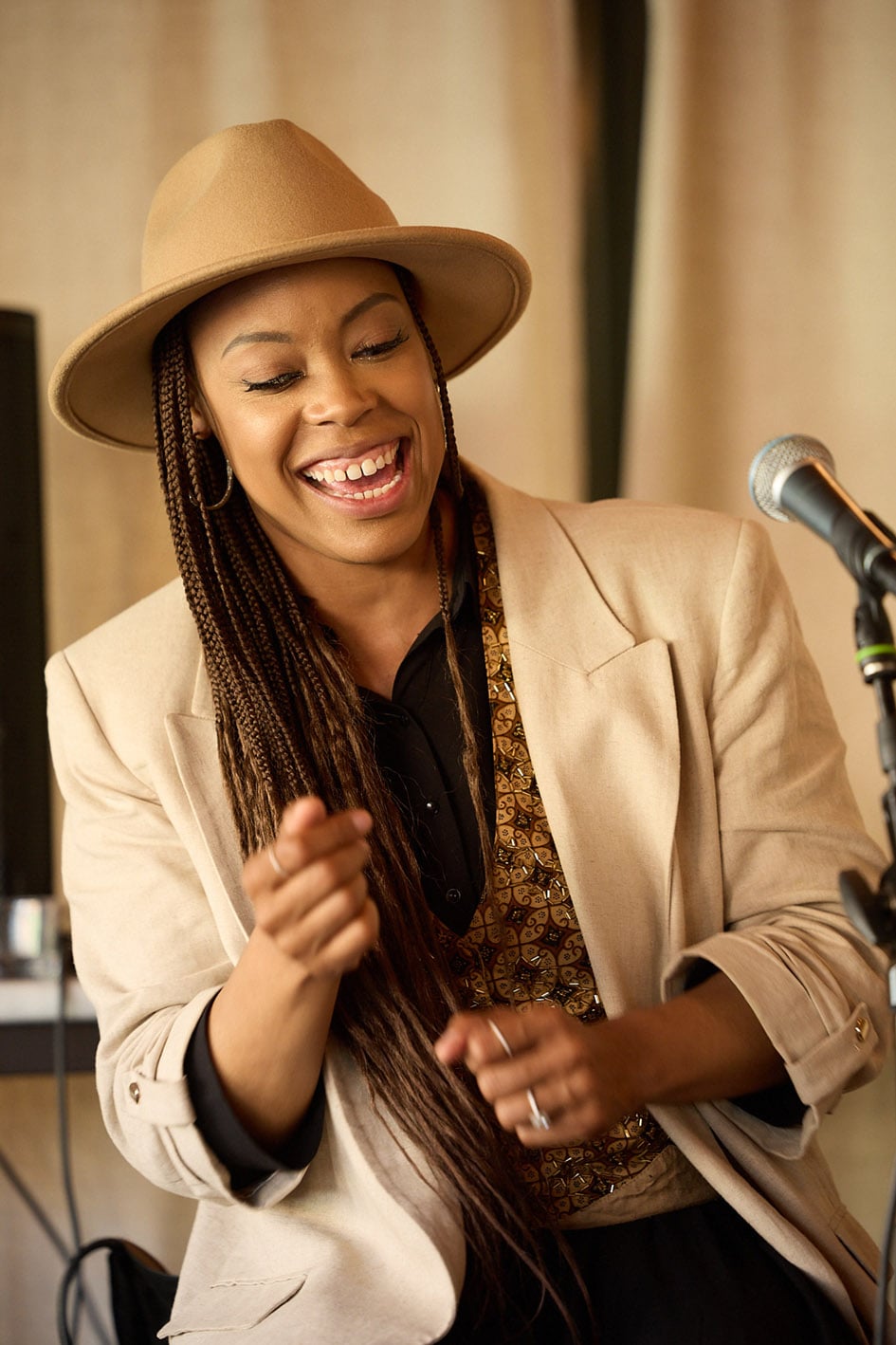 Kiralina sitting and singing in a mic wearing a light brown hat vest and cream blazer.