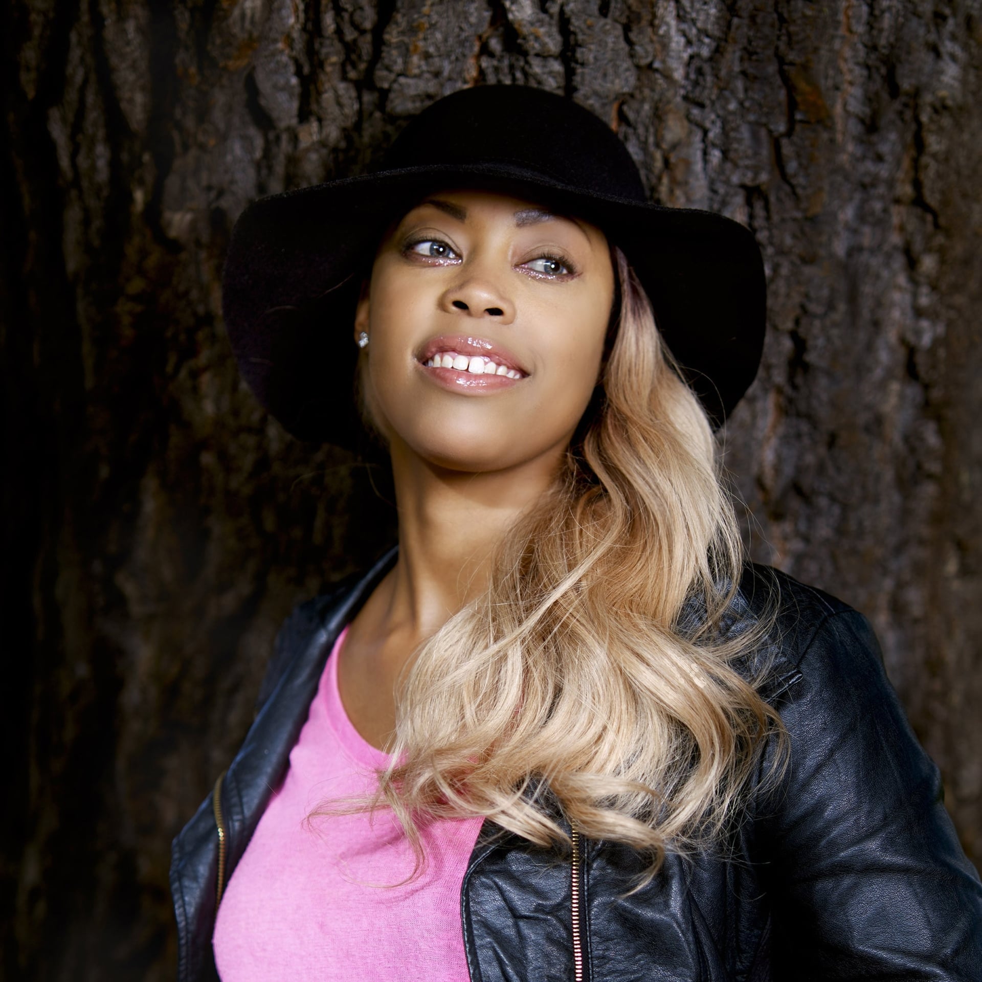 Kiralina is leaning on a tree with black hat and a pink t-shirt and black leather jacket.